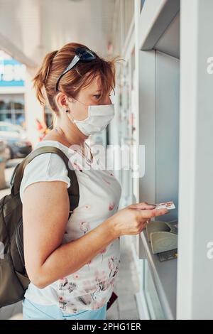 Frau, die Geld vom geldautomaten mit Debit- oder Kreditkarte abhebt, steht draußen auf der Straße und trägt eine Gesichtsmaske, um dabei Mund und Nase zu bedecken Stockfoto