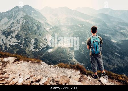 Junger Mann mit Rucksackwandern in einem Berg, der aktiv Sommerurlaub verbringt. Rückansicht eines Mannes, der auf einem Hügel steht und die Berge von ian bewundert Stockfoto