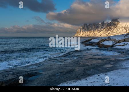 Am Fels sich brechende Wellen des Atlantik bei Tungeneset, Senja, Norwegen, mit den steilen Bergen Okshornan im Hintergrund. Stürmisches Winterwetter Stockfoto