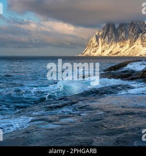 Am Fels sich brechende Wellen des Atlantik bei Tungeneset, Senja, Norwegen, mit den steilen Bergen Okshornan im Hintergrund. Stürmisches Winterwetter Stockfoto