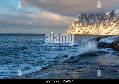 Am Fels sich brechende Wellen des Atlantik bei Tungeneset, Senja, Norwegen, mit den steilen Bergen Okshornan im Hintergrund. Stürmisches Winterwetter Stockfoto