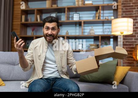 Porträt eines glücklichen Online-Käufers zu Hause, eines erwachsenen Mannes, der lächelt und in die Kamera schaut, die Produktverpackung und das Telefon in der Hand hält und sich über die schnelle Lieferung aus dem Online-Shop freut. Stockfoto