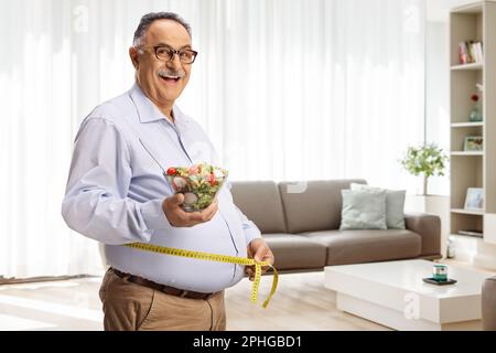 Ein erwachsener Mann hält einen Salat und misst die Taille in einem Wohnzimmer zu Hause Stockfoto