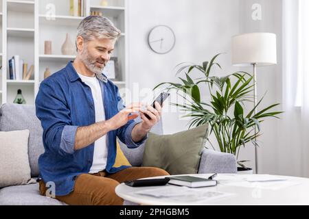 Ein grauhaariger Mann, der zu Hause auf dem Sofa sitzt und telefoniert. Stockfoto