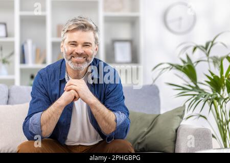 Porträt eines gruseligen, grauen Mannes in einem blauen Hemd, der zu Hause auf der Couch sitzt, in die Kamera lächelt und seine Arme vor ihm kreuzt. Stockfoto