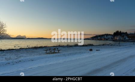 Abendrot mit Brücke, Rastplatz und Baum; Panorama, Verschneite Berge in Senja, Norwegen. roten Wolken spiegeln sich in Wasser des Fjords bei Skaland Stockfoto