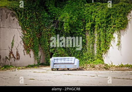 Altes verlassenes Sofa in einer Ecke des Betonparkplatzes mit hohen Betonwänden im Hintergrund, die mit Reben bedeckt sind - sieht aus wie ein Fotoshooting-Set Stockfoto