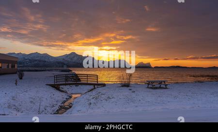 Abendrot mit Brücke, Rastplatz und Baum; Panorama, Verschneite Berge in Senja, Norwegen. roten Wolken spiegeln sich in Wasser des Fjords bei Skaland Stockfoto