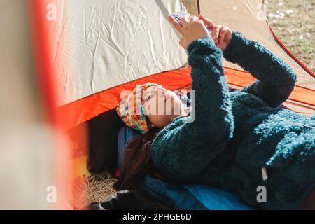 Junge Reisende Frau, die warme Kleidung trägt, ihr Handy benutzt, das morgens in einem Campingzelt liegt. Stockfoto