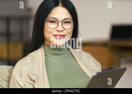 Eine fröhliche Asiatin mittleren Alters mit Brille surft gern auf ihrem Tablet im Internet, während sie bequem zu Hause sitzt. Hochwertiges Foto Stockfoto