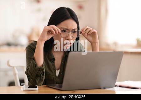 Schockierte asiatische Frau, die am Schreibtisch sitzt und auf den Laptop schaut Stockfoto