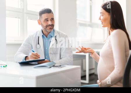 Junge schwangere Frau, die im Krankenhaus sitzt, spricht und sich mit einem männlichen Gynäkologen mittleren Alters beraten hat Stockfoto