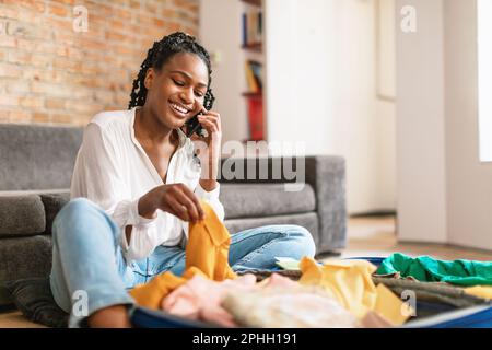 Positive schwarze Frau, die Kleider in den Koffer packt und mit dem Handy spricht, die Reisetour telefonisch von zu Hause aus gebucht hat Stockfoto