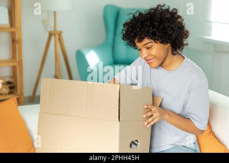 Afrikanisches Mädchen, das die Lieferung auspackt und in die Schachtel schaut. Glückliche Frau, die eine Schachtel öffnet. Eine Frau bekommt ein Paket, das sich die gelieferten Waren ansieht. Zufriedene Kunden positives Feedback, schnelle Expresslieferung Stockfoto
