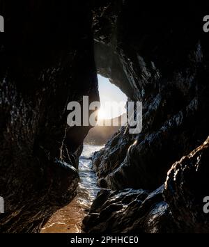 Eine Meereshöhle bei Sonnenuntergang durch die Felsen der Kynance Cove auf der Lizard Peninsula, Cornwall, wenn die Sonne untergeht und die Flut kommt. Stockfoto