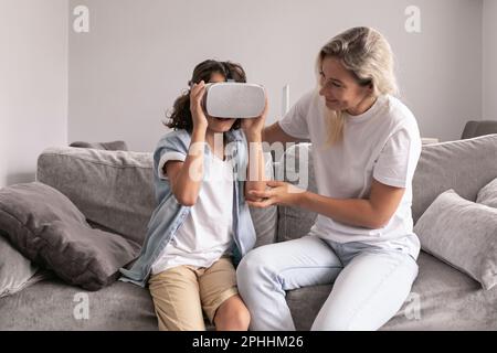 Mutter hilft seinem Sohn, im Wohnzimmer zu Hause eine Virtual-Reality-Headset-Brille zu tragen und Spaß zu haben Stockfoto