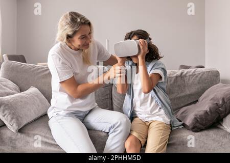 Mutter hilft seinem Sohn, im Wohnzimmer zu Hause eine Virtual-Reality-Headset-Brille zu tragen und Spaß zu haben Stockfoto