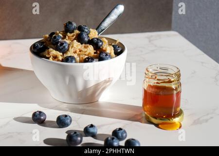 Gesunder Haferbrei mit Blaubeeren oben in weißer Porzellanschüssel, Honigdose und Orangen im Hintergrund Stockfoto