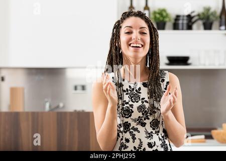 Junge, schöne Frau klatscht in ihrer atemberaubenden Küche. Sie applaudiert dafür, die Situation zu unterstützen und zu engagieren, zuzustimmen und zu genießen. Mit geflochtenem Haar und Stockfoto
