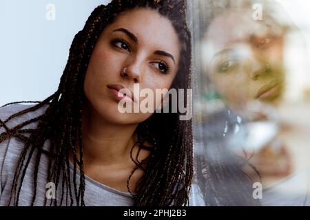 Nahaufnahme einer jungen Frau, die aus dem Fenster schaut, reflektiert in einem Farbeffekt. Sie ist seriös und aufmerksam, intelligent, mit Boxgeflechten, nein Stockfoto