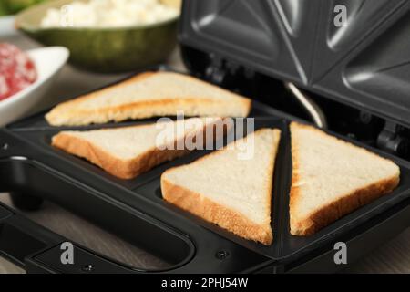 Moderner Sandwichautomat mit Brotscheiben auf Holztisch, Nahaufnahme Stockfoto