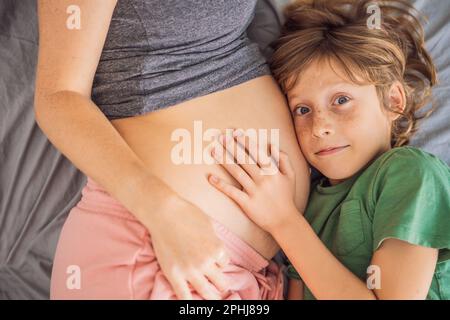 Junge, schöne, schwangere Frau und ältester Sohn. Der süße Junge lehnte sein Ohr gegen den Bauch seiner Mutter. Ein Baby im Familienkonzept erwarten Stockfoto