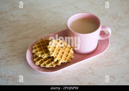 Ein rosafarbenes Set aus einer Tasse und einem Teller mit hausgemachten Waffeln und Cappuccino zum Frühstück am frühen Morgen an einem sonnenbeleuchteten Tisch. Stockfoto