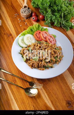 Gebratene Nudeln oder Mie Goreng mit Gemüse, Huhn, Ei und Rindfleisch auf weißem Teller am Holztisch umrühren Stockfoto