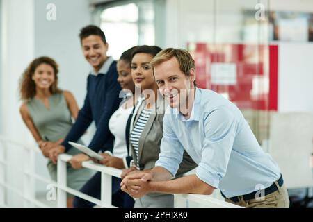 Dieses Team strebt Erfolg an. Porträt einer Gruppe lächelnder Mitarbeiter, die sich in einem Büro auf ein Geländer lehnen. Stockfoto