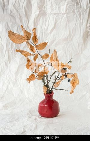 Stillleben Herbstblüten Vase. Retro-Style. Getrocknete orangefarbene Buchenblätter stehen in einer dekorativen Vase vor einem Hintergrund aus zerknittertem weißem Papier. Der Betrug Stockfoto