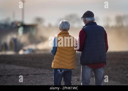 Rückansicht von zwei Bauern, Mann und Frau, die auf dem Feld gehen, während der Traktor im Hintergrund gepflügt wird Stockfoto