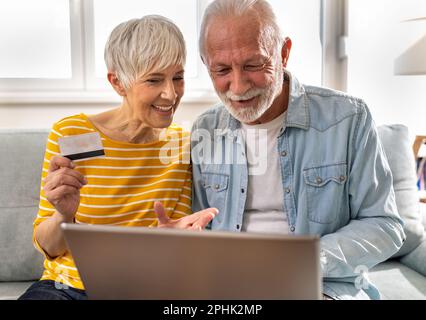 Aufgeregtes Seniorenpaar, das online auf einem Laptop einkauft, auf dem Sofa im Wohnzimmer sitzt Stockfoto