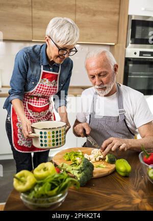Seniorenpaar bereitet Suppe zu, Ehemann schneidet Gemüse, während Frau Pot hält Stockfoto