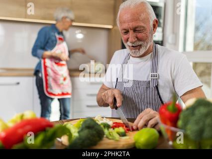 Seniorenpaar bereitet Suppe zu, Ehemann schneidet Gemüse, während Ehefrau im Hintergrund kocht Stockfoto