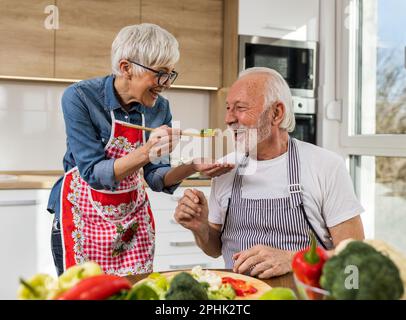 Ein fröhliches Seniorenpaar, das Essen zubereitet, ein Mann, der gekochtes Gemüse aus der Schaufel probiert Stockfoto