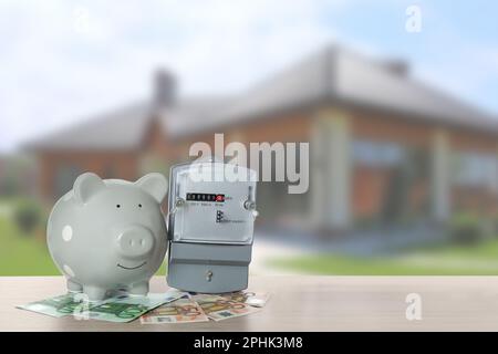 Stromzähler, Sparschwein und Geld auf Holztisch gegen verschwommene Sicht auf das moderne Haus. Platz für Text Stockfoto
