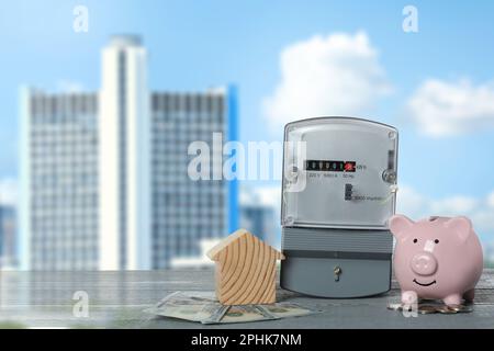 Stromzähler, Hausmodell, Geld und Sparschwein auf grauem Holztisch gegen verschwommene Sicht auf die Stadt Stockfoto