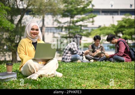 Attraktive und glückliche junge asiatische muslimische Studentin im Hijab, die auf dem Gras sitzt und ihren Laptop im Campuspark benutzt. Stockfoto