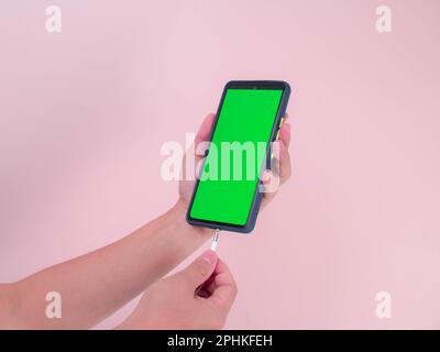 Weibliche Hände stecken den Kopfhörer in das Smartphone mit grünem Bildschirm auf pinkfarbenem Hintergrund. Nahaufnahme des Handeinsteckens von Kopfhörern in Smartp Stockfoto