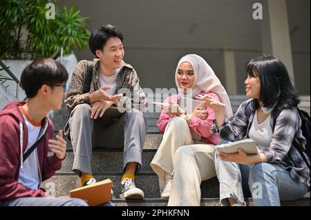 Eine Gruppe von asiatisch-vielfältigen Studenten unterhalten sich und entspannen sich auf den Treppen außerhalb des Campus-Gebäudes zusammen. Freundschaft und Bildung Betrug Stockfoto