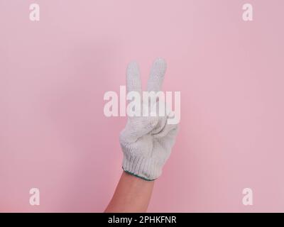Hände in weißen Strickhandschuhen isoliert auf pinkfarbenem Hintergrund. Weibliche Hand, die mit einem Baumwollhandschuh zwei Finger hochhält (Friedenssymbol). Stockfoto