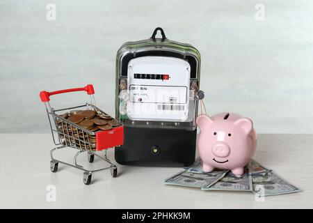 Stromzähler, Sparschwein, kleiner Einkaufswagen mit Münzen und Dollarscheine auf einem weißen Holztisch Stockfoto