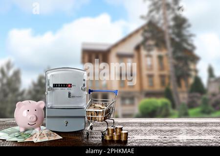 Stromzähler, Sparschwein und kleiner Einkaufswagen mit Geld auf Holztisch gegen verschwommene Sicht auf das wunderschöne Haus Stockfoto