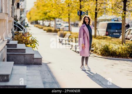 Frau Herbststadt. Eine Frau in einem rosa Kunstpelzmantel, die im Herbst an einem sonnigen Tag auf einer Straße posiert. Bäume mit gelben Blättern entlang der Straße Stockfoto