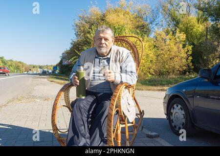 Porträt eines ukrainischen Seniors, der am Straßenrand in einem Schaukelstuhl sitzt und eine Vakuumflasche hält, die sich am sonnigen Herbsttag für Tee bereitstellt Stockfoto