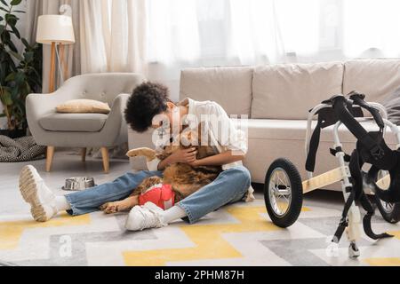 Junge afroamerikanische Frau umarmt behinderten Hund in der Nähe eines Rollstuhls im Wohnzimmer, Stockbild Stockfoto