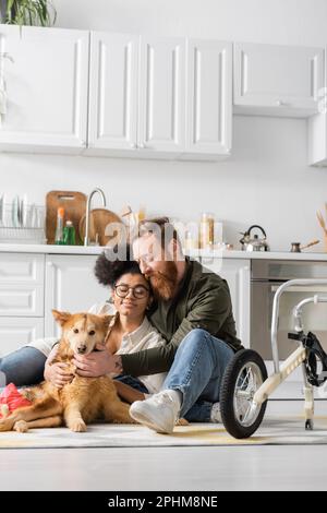 Multiethnisches Paar, das in der Nähe eines behinderten Hundes auf einem Teppich in der Küche sitzt, in der Nähe eines Rollstuhls, Stockbild Stockfoto