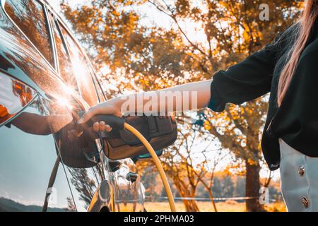 Weibliche Hand, die ein Ladegerät in eine geöffnete Ladebuchse eines schwarzen Elektroautos einsteckt, wobei die Sonne durch einen grünen Baumkamm im Hintergrund scheint Stockfoto