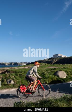 Ein Radfahrer mit Schutzhelm, der auf einem roten Fahrrad bergauf auf der Küstenstraße mit Blick auf Newquay Bay in Cornwall im Vereinigten Königreich fährt. Stockfoto