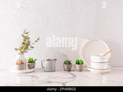 Weiße Keramikutensilien und Innenblumen in Keramik-Minitöpfen auf der Küchenarbeitsfläche aus weißem Marmor. Graue Zementwand. Ein Kopierbereich Stockfoto
